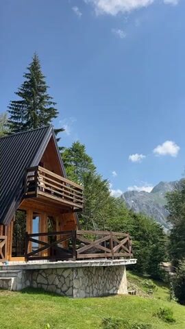A wooden cabin with a black roof sits on a stone foundation surrounded by green grass. A wooden balcony extends from the upper level of the cabin, and a person in a white top and shorts is visible on the balcony. Another wooden railing is attached to the lower part of the cabin. The cabin is nestled among lush green trees, with a tall evergreen tree to the left. In the background, majestic mountains are visible under a clear blue sky with scattered white clouds. A dirt path winds its way up the grassy slope to the right of the cabin.