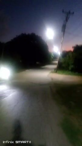 The video shows a first-person perspective of someone moving down a dark road at night, illuminated by streetlights. The road is surrounded by trees and vegetation, creating a silhouette against the twilight sky. The camera shakes slightly as the person moves. The words "Infinix SMART 8" appear at the bottom of the screen.