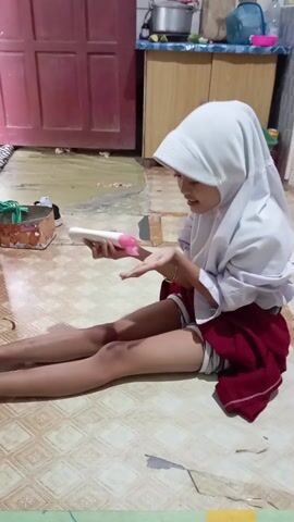 A young girl in a white headscarf, a white shirt, and a red skirt sits cross-legged on a tiled floor indoors. She holds a white bottle in her left hand and is squeezing a pink lotion or cream onto her right palm. Behind her, there is a red door, a wooden cabinet, and some household items. To her left, there is a container on the floor. The lighting is soft, and the scene appears to be casual and domestic.