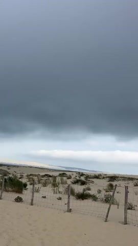 The video shows a sand dune landscape under a cloudy sky. A wire fence with wooden posts runs across the foreground, with sparse vegetation growing in the sand behind it. In the middle ground, a family walks along a sandy path. A man wearing a blue shirt and shorts carries a child in a backpack carrier. Beside him, a woman in a patterned shirt and white shorts walks with him. Further down the path, another woman in a white shirt and black shorts walks away from the camera, carrying a bag. In the background, a long ridge of sand dunes stretches across the horizon, with more distant hills visible. The sky is filled with heavy, dark gray clouds, suggesting an overcast or stormy day.