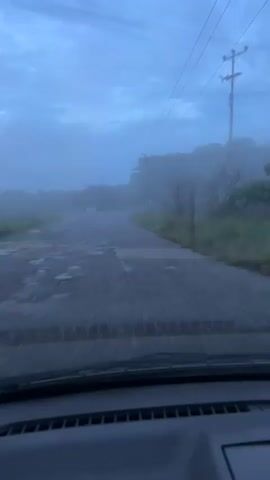 A point-of-view shot from inside a moving vehicle captures a wet, cracked paved road on an overcast and foggy day. The vehicle drives forward, revealing a fork in the road where one path veers to the right. Power lines, utility poles, and trees line the sides of the road, partially obscured by the low visibility. A blue road sign is visible on the right side near the diverging path. The vehicle's dashboard is partially visible at the bottom of the frame.