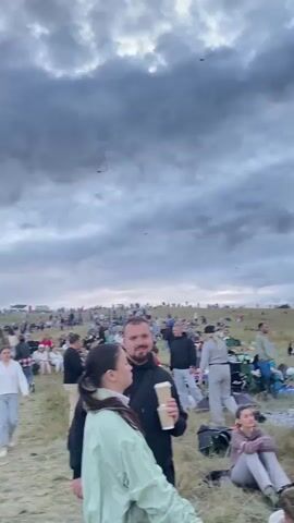 The video shows a wide outdoor scene with a large crowd of people gathered on a grassy hill under a cloudy sky. Many people are sitting on blankets and in lawn chairs, appearing to be attending an event. In the sky, several kites are visible. The camera pans across the crowd and the landscape, showing groups of people interacting and observing the surroundings. There are trees and a distant structure visible in the background. The overall atmosphere appears relaxed and communal, with people enjoying the outdoors and the kite-flying activity.
