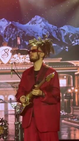 The media file captures a live musical performance. The main subject is a male guitarist with distinctive, spiky dreadlocks, wearing oversized red sunglasses and a matching red suit jacket and pants. He is playing a light-colored electric guitar, which has a dark strap with a patterned design.

The setting appears to be a stage with a large backdrop depicting a snowy mountain range under a starry night sky. In front of the mountains, there is a stylized architectural element that resembles a grand building with ornate details and what looks like a large emblem featuring an "S" inside a shield. Stage lighting, including a bright spotlight from the upper left, illuminates the performer.

To the left of the guitarist, drum equipment is visible, including a snare drum and what appears to be a Fender Twin Reverb amplifier. A microphone on a stand is positioned near the guitarist.

The overall impression is of a concert or a performance event. The performer's attire and hairstyle suggest a modern, possibly artistic or alternative style. The backdrop creates a dramatic and somewhat fantastical atmosphere. There is no discernible weather or specific time of day beyond the depiction of night on the backdrop. The location context indicates that this event is taking place in Estosadok, Russia. The visible text on the amplifier reads "FENDER TWIN REVERB."
