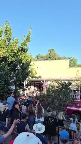 The video shows a street lined with people watching a parade. There are buildings on either side of the street, with some decorated with American flags. A red fire truck is visible in the foreground, and a few trees are present. The sky is clear and blue. The people are standing and sitting along the street, some are holding up phones to record the parade.