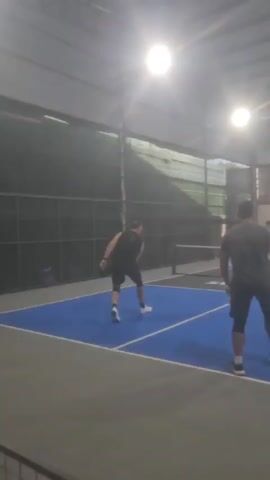 Four people are playing pickleball indoors on a blue court with gray borders. The court is enclosed by dark fencing and a metal roof with overhead lighting. Two men, one wearing a gray shirt and black shorts, and another in a black tank top and black shorts, are on one side of the net, positioned closer to the camera. The man in the gray shirt is mid-swing with a paddle. On the other side of the net, two other men, one in a blue shirt and black shorts and another in a dark shirt and black shorts, are also positioned on the court. The man in the blue shirt is closer to the net, and the man in the dark shirt is further back. The game appears to be in progress.