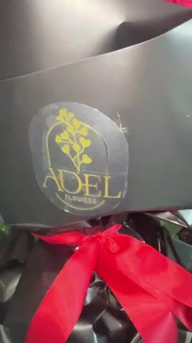 The video shows a bouquet of red roses in a black paper wrapping. The bouquet has a red ribbon tied around the bottom. The wrapping has a sticker with the name "ADEL FLOWERS" on it. The camera moves slightly, showing the roses from different angles. The background is dark.