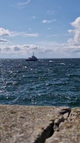A choppy, dark blue sea under a bright sky with scattered white clouds. In the distance, a tugboat with a tall mast is sailing. To the left of the tugboat, a kite surfer is visible. The foreground of the image is blurred, showing a rough, grey concrete or stone surface. The overall impression is a windy day at sea.