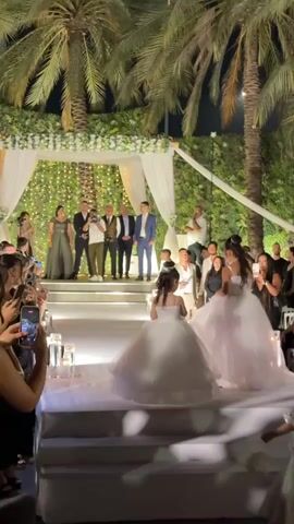 This is a video of a wedding ceremony taking place outdoors at night. In the foreground, two young girls in white dresses are walking down an aisle. In the background, a wedding party stands under a chuppah decorated with lights and flowers. Palm trees are visible on either side of the chuppah. Guests are seated on either side of the aisle, and some are taking pictures with their phones. The overall atmosphere appears festive and celebratory.