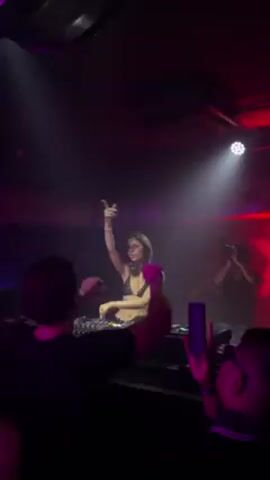 The video shows a DJ, a woman with dark hair, performing in front of a crowd in what appears to be a nightclub or concert venue. She is standing behind a DJ console, with her hands up in the air as if encouraging the crowd. The lighting is dark with red and white spotlights highlighting the DJ and a large red "X" logo behind her. People in the crowd are holding up their phones to record the performance.