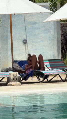 A woman in a polka dot bikini and sunglasses lies on a colorful chaise lounge by a pool. She has her legs raised and crossed, and her hair is in a bun. There's an umbrella over her head and a shower stall behind her. Other beach chairs are on the pool deck. The pool water is slightly rippled.