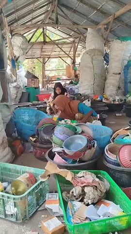 The video shows two women sorting through piles of plastic waste in a makeshift open-air shelter. The shelter has a corrugated metal roof supported by wooden beams. The floor is dirt and scattered with debris. In the foreground, there are green plastic crates filled with various recyclable materials, including cans and paper. Behind the crates are large piles of sorted plastic items, such as bowls, basins, and buckets in various colors and sizes, as well as some larger, blue plastic containers. The two women are squatting amidst these piles. The woman on the right, wearing a blue shirt and an orange headscarf, is actively handling plastic items and appears to be sorting them. The woman on the left, wearing a black headscarf and a patterned shirt, is also engaged in sorting. In the background, there are more large, white sacks, possibly filled with compressed recyclable materials, and other discarded items. The overall impression is of a recycling or waste sorting operation in a rural or informal setting.
