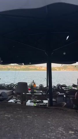 A slow-motion, low-angle, wide shot shows a restaurant patio overlooking a body of water. A large, black umbrella covers the patio, and tables are set with white tablecloths, green placemats, and bottles of wine. Several people are seated at the tables, and a lamp is visible on one of the tables. The water is calm, and a distant shore with buildings is visible. The overall atmosphere is serene and relaxing.