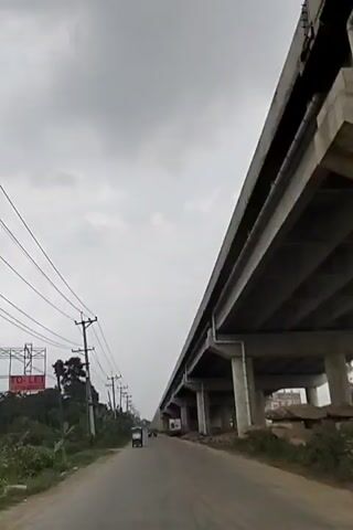 The video shows a low-angle view of a road beneath a large, elevated concrete structure, likely a highway or train track. The sky is overcast and gray. On the left side of the road are power lines and vegetation.  A black car drives towards the camera. Other vehicles, including what appears to be a small, three-wheeled vehicle, are visible further down the road. Debris and construction materials are visible on the right side of the road. The overall impression is of a somewhat rural or developing area with modern infrastructure being constructed.