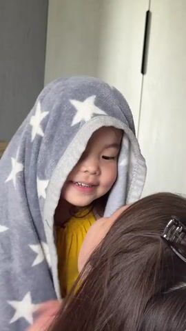 A toddler wearing a yellow dress and wrapped in a gray blanket with white stars hides behind an adult woman's head before peeking out and looking at the camera. The adult woman has dark brown hair with a black hair clip in the back. The scene takes place indoors, with a gray wall and a light brown wooden stool in the background.