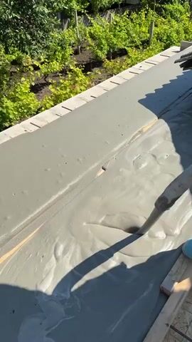 A concrete hose is being used to pour concrete into a formwork. The concrete is wet and fluid, creating ripples and swirls as it is poured. The formwork appears to be on a rooftop, with greenery visible in the background. There are wooden planks and other construction materials around the formwork. A person's legs, wearing blue shoes or boots, are visible near the hose. The sun is shining, casting shadows on the concrete.