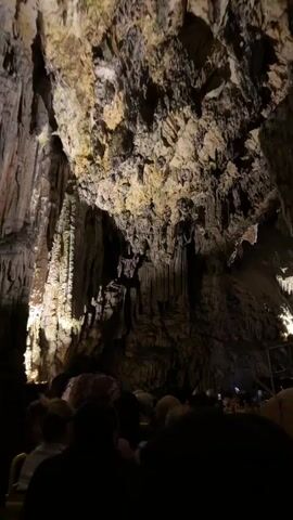 The video displays the interior of a vast, dimly lit cave, filmed from the perspective of a passenger on a slow-moving vehicle. The cave's ceiling and walls are extensively covered with stalactites and other natural rock formations, highlighted by strategically placed artificial yellowish lights. The camera pans slowly, showcasing these geological structures. In the foreground, the backs of multiple seated individuals are visible, observing the cave. One person is seen holding a smartphone aloft, actively recording the surroundings. The vehicle continues to move through the cavernous environment.