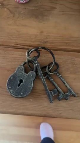 The video shows a close-up, top-down view of a set of antique-looking keys and a padlock, resting on a wooden surface. The keys are made of dark metal with a distressed finish, and they are attached to a large metal ring. There are three keys, each with a different intricate keybit. The padlock is also dark metal with a distressed finish and features a keyhole. The wooden surface appears to be a table or shelf with visible grain. At the very bottom of the frame, a small portion of a person's foot, wearing a pink sock or shoe, is visible. The lighting is dim, and the focus is sharp on the keys and padlock. The camera angle is stable, and there is no visible movement from the keys or padlock themselves.