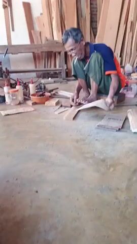 An elderly man wearing glasses, a blue and orange t-shirt, and green pants is sitting on the floor in a workshop, crafting a piece of wood. He is barefoot and is using a tool to shape the wood, with a cigarette in his mouth. Various pieces of wood are stacked behind him, and on the floor around him are tools, bottles of glue, and other materials. The workshop appears to be well-lit and filled with lumber.