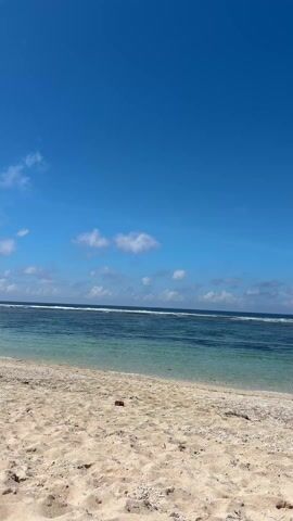 The video shows a sunny beach scene. The foreground is a sandy beach with some pebbles and debris. The midground features a body of water with turquoise near the shore transitioning to a deeper blue further out, where gentle waves are visible. The background consists of a clear, bright blue sky with scattered white clouds. There are no people visible in the video. The camera is static.