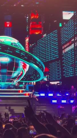 A concert is taking place on a stage. The stage is decorated with a large, circular, green and blue light installation that resembles a globe. Behind the globe is a large screen displaying a cityscape with numerous neon lights and buildings. A man in a dark shirt and pants is standing center stage, facing the audience, with his arms raised. Two other individuals, presumably band members, are standing on either side of him, holding guitars. A drum set is visible on the right side of the stage. The audience is visible in the foreground, with many people holding up their phones to record the performance. The overall atmosphere is energetic and vibrant, with flashing lights and a concert atmosphere.