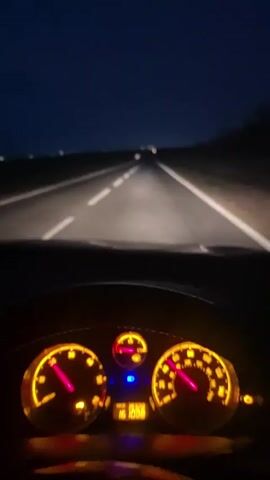 A view from the driver's seat shows a car driving at night on a straight road with double yellow lines. The instrument panel is visible, illuminated with yellow lights. The speedometer reads around 60 mph, and the tachometer reads around 2500 RPM. The exterior is dark. There are other lights visible in the distance.  Music is playing.