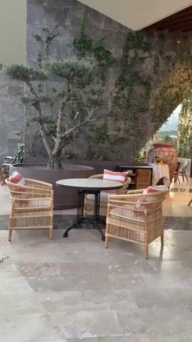 The video pans across an outdoor seating area of a restaurant or hotel. In the foreground, there are two wicker chairs with striped cushions and a round table with a dark metal base. Behind these, a large potted olive tree with a gnarled trunk is visible against a stone wall covered in ivy. Further back, more tables and chairs are arranged, some with wooden tops and woven seats. Beyond the seating area, a mountainous landscape with green trees is visible under a bright sky, suggesting daytime. The overall atmosphere appears relaxed and inviting.