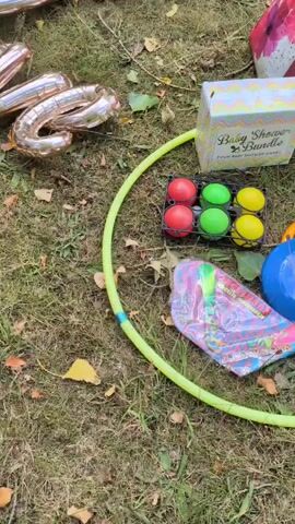 The video pans over a patch of grass with fallen leaves. In the foreground, there are rose gold foil balloons spelling out "LOVE". To the left, a bright yellow hula hoop lies on the grass. To the right of the balloons, there is a box labeled "Baby Shower Bundle" with six colorful balls inside a black holder. Further to the right, a blue frisbee is visible. In the background, a red bag and some silver poles are partially visible, suggesting an outdoor setting.