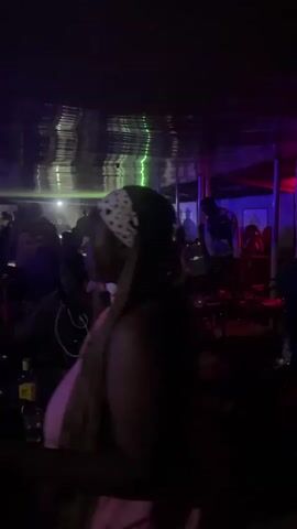This media file captures a vibrant scene inside a dimly lit establishment, likely a club or lounge, in Durumi, Nigeria. The atmosphere is electric, illuminated by a mix of purple and green lights that create a dynamic, almost psychedelic ambiance. The ceiling reflects the colorful lighting, adding to the immersive visual experience.

The primary focus is on a group of people socializing and enjoying themselves. In the foreground, a woman with her back to the camera is visible, her braided hair cascading down her shoulders, adorned with a bandana. To her right, another woman in a white dress with a floral pattern is engaged in conversation and appears to be laughing, her face alight with joy. Other individuals are scattered throughout the space, some seated, others standing, all contributing to the lively social gathering.

The setting suggests an indoor entertainment venue, possibly a shisha lounge given the presence of what appear to be shisha pipes on tables. The overall mood is one of celebration and camaraderie. The time of day is clearly nighttime, as evidenced by the artificial lighting and the dark surroundings. There are no discernible weather cues or outdoor elements visible.

No visible text is present in the media file. The scene is characterized by the energy of the people, the pulsating lights, and the general aura of a night out in Durumi, Nigeria.