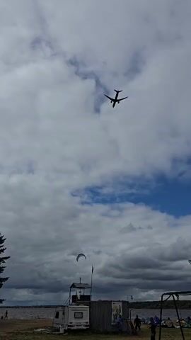 A low-angle shot captures a cloudy sky with an airplane flying overhead. Below, a waterfront scene unfolds with a small building and a camper van. In the distance, a body of water is visible, and on the shore, a few people and tents suggest an outdoor gathering. The airplane is positioned in the upper portion of the frame, appearing as a dark silhouette against the sky. A kite is also visible in the sky, to the left of the small building. The overall atmosphere is one of a casual outdoor event on a day with mixed weather conditions.