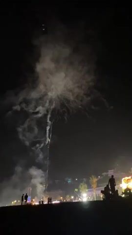 The video captures a nighttime fireworks display over a coastal town. Silhouettes of people are visible in the foreground, gathered on what appears to be a beach or elevated viewpoint. In the background, a town with illuminated buildings and palm trees is spread out along the coastline, with a hillside dotted with more lights. Fireworks burst in vibrant green and golden hues against the dark sky, leaving trails of smoke. The overall atmosphere suggests a festive event.