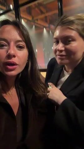 Two women are sitting next to each other in what appears to be an indoor restaurant or lounge. The woman on the left with long brown hair is holding the camera, filming herself and her friend who is on the right. Both women are smiling and engaging with the camera. The woman on the right is wearing a black blazer, a white shirt, and a gold bracelet. The woman on the left is wearing a brown top and a necklace. There are wicker chairs and a glass window with reflections of the outside behind them. The woman on the left says "You know guys, we are having a really really good time".