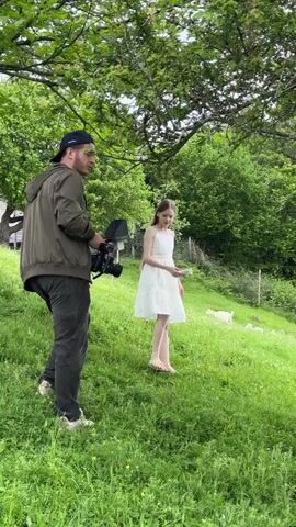 A man with a camera stands on a grassy hill, watching a young girl in a white dress walk towards him. The man wears a green jacket, dark pants, a baseball cap, and light-colored sneakers. He is holding a professional-looking camera. A sprig of greenery is tucked behind his ear. The girl has long hair and is barefoot. Behind her, in the background, a few white goats are grazing in the field. The scene is lush and green, with trees and foliage surrounding the clearing.