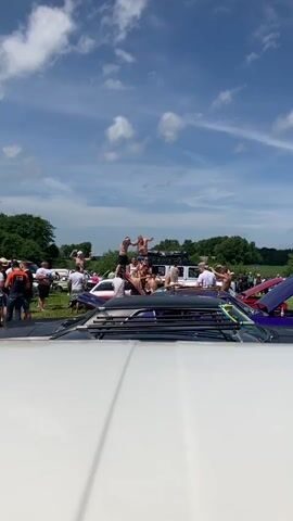 The video shows an outdoor gathering of people on a sunny day. Several individuals are standing or sitting on the roofs and hoods of cars, some of which appear to be classic models. The people are casually dressed, many in swimwear. Some are holding drinks and gesturing or dancing. The environment appears to be a field with trees in the background and a blue sky with scattered clouds. The camera perspective is from the front of a white vehicle, looking over its hood towards the gathering. The video appears to be a short, unedited clip of the event.