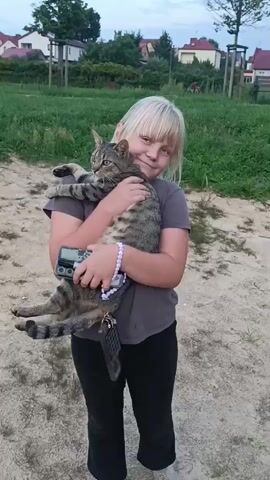 A young girl with blonde hair and a grey t-shirt is holding a tabby cat. She is wearing black pants and white shoes. The cat is looking at the camera. The girl is smiling. She is holding a small electronic device in her left arm and another one hanging from her wrist. The background is a park with trees and houses. The ground is dirt and gravel.