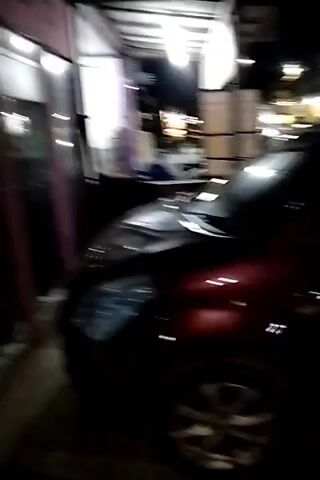 The video begins with a shot of the side of a dark red car parked at night. The car's tires and side-view mirror are visible. The camera then pans to the right, showing the interior of a dimly lit store. The store has tiled floors and pink framing around the entrance. Inside, there is a chair, what appears to be display shelves with items on them, and decorative elements on the wall. The store's lights are on, illuminating the space. The video is shaky and appears to be taken with a mobile phone.