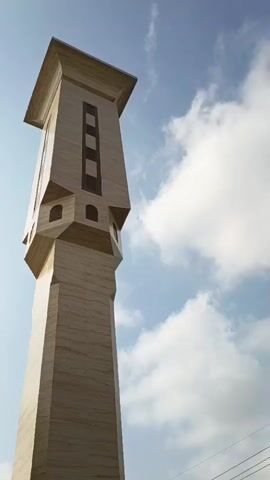 This is a low-angle, upward-looking video of a tall, modern minaret against a blue sky with scattered clouds. The minaret is constructed from light-colored, veined stone. It has a complex, multi-tiered design. The base is a tall, octagonal shaft. Above this, there is a section that flares outwards with angular projections, each featuring small, arched openings. Higher still, the minaret narrows and features a rectangular section with three dark, vertical panels. The uppermost part of the minaret is a square cap with a slightly overhanging roof. The video rotates very slowly, showing the minaret from slightly different angles.