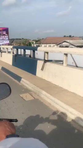 The video shows a view from what appears to be a moving vehicle, likely a motorcycle or similar conveyance, traversing a bridge. The bridge has a concrete barrier, painted in white and dark blue, along its side. Visible beyond the barrier are buildings, trees, and grass. A large billboard promoting "Halifield School College, Port Harcourt" is prominent on the left. The sky is partially cloudy. The road surface and curb are also visible. The camera movement indicates the vehicle is moving forward along the bridge.