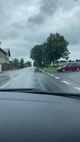 The video is shot from inside a car, looking out the windshield which has raindrops on it. The car is on a wet, paved road, indicating it is raining or has recently rained. The road appears to be in a rural or suburban area, with houses and trees lining the side. There are street signs and traffic signs visible. A red car is parked on the side of the road to the right. The sky is overcast with grey clouds. The video shows the car moving forward along the road.