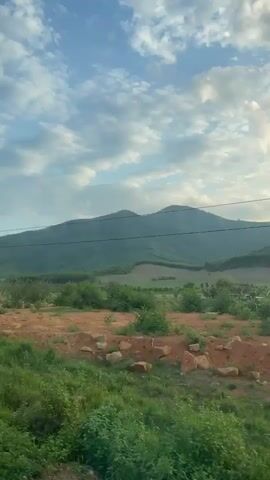 A daytime, eye-level shot captures a moving view of a rural landscape. In the background, two prominent, tree-covered mountains rise under a partly cloudy blue sky with scattered white clouds. The middle ground consists of diverse terrain featuring patches of green grass, reddish-brown earth, and scattered green bushes or small trees, with what appears to be cultivated fields or plantations on the lower mountain slopes. The immediate foreground is dominated by vibrant green grass interspersed with some dry patches. A thin, dark utility wire spans horizontally across the upper portion of the frame. The camera moves smoothly forward or pans gently to the right, continuously revealing the passing scenery.