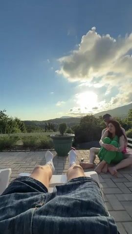 The video opens with a close-up view of a person's legs, clad in denim shorts and white socks, resting on a white lounge chair. The perspective is from the viewpoint of someone lying down. The camera then pans out slightly to reveal a picturesque outdoor setting.

In the mid-ground, two individuals are seated on a paved patio area. One person, dressed in a pink top and green pants, is being embraced from behind by another person wearing a dark cap. They appear to be a couple, sitting close together and looking towards the left of the frame. The woman is resting her head on the man's chest.

The background features a lush, green landscape with rolling hills and trees. A large potted plant with a small tree inside sits prominently on the patio. The sky is a vibrant blue, with dramatic, fluffy white clouds catching the warm glow of the setting sun, which is partially obscured by the clouds and the distant mountains. The overall impression is of a serene and romantic evening.

The time of day is clearly late afternoon or early evening, suggested by the low angle of the sun and the warm, golden light bathing the scene. The weather appears to be pleasant and clear, with the sun shining through.

The location context is identified as Shirokaya Balka, Russia, which suggests a potentially coastal or mountainous region known for its natural beauty.

There is no visible text in the video. The mood is peaceful and relaxed, with a sense of intimacy and shared enjoyment of the beautiful scenery and sunset.
