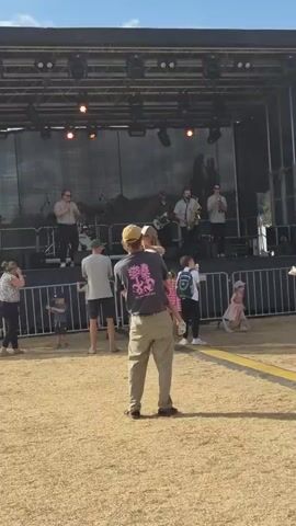 A man in khaki pants and a grey t-shirt with a pink graphic on the back holds a young child in his arms. The man wears a tan baseball cap. The child is wearing a pink dress and has a blonde ponytail. They are standing on a dry grassy field facing a stage. On the stage, a band is performing. The band consists of at least five members playing various instruments including a saxophone, a trumpet, a guitar, and drums. The stage is set up outdoors with a black backdrop and a light rig above. A metal barrier separates the audience from the stage. Other people, including children, are visible in the foreground and background, some standing and some sitting. The sky above is blue with some wispy clouds.
