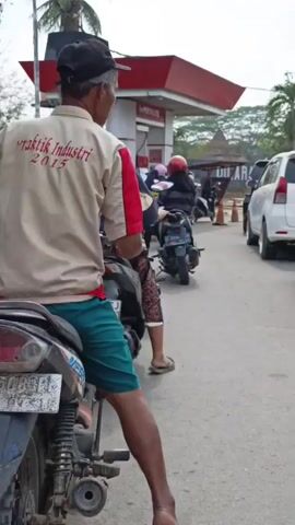 A man wearing a tan shirt with "Praktik Industri 2015" printed on it and green shorts sits on a motorcycle. He is looking to his right. Behind him, a woman wearing a black hijab and a patterned skirt is also sitting on the motorcycle. The motorcycle is stopped in a line of vehicles at a gas station. The gas station has a red roof and a sign that reads "PERTAMINA." There are other motorcycles and cars in the line. The ground is paved. The lighting indicates it is daytime.