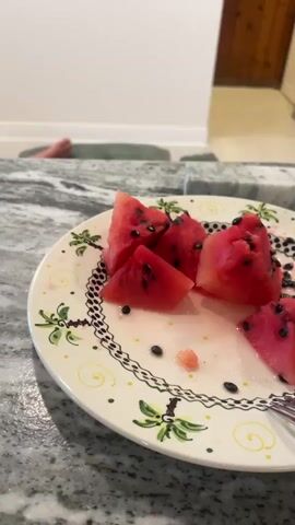 A close-up shot on a plate of watermelon slices is followed by a wider view of a living room where a large television is displaying a soccer game. The camera focuses on the television screen, which shows players on a green field with yellow-clad spectators in the background. In the foreground, the edge of a gray countertop is visible, and to the right, a wooden paneled wall section is present. A portion of a person's bare foot and leg are visible on a green, ribbed cushion in front of the television, suggesting someone is relaxing and watching the game. The room appears to be well-lit, with a white wall and baseboard.