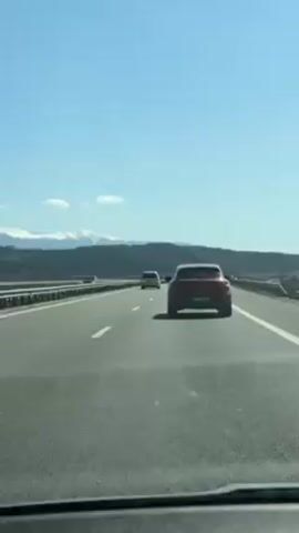 The video shows a view from the passenger seat of a car driving on a highway.  The sky is clear and blue with a few white clouds.  In the distance, a mountain range covered in snow is visible.  Several other cars are on the highway, including a red SUV in front of the camera car. The road is asphalt and has white lane markings. A metal guardrail separates the highway from the landscape. The overall impression is one of a sunny day and pleasant travel.