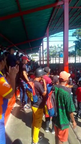 The video shows a crowd of people walking into a stadium seating area. The area is covered by a green metal roof supported by red pillars. The crowd is diverse in terms of clothing, and many are wearing hats and carrying backpacks. A chain-link fence separates the walking area from the stadium seats, which are mostly white and blue plastic. There is a multi-colored banner hanging in the background. The video is shot from within the covered area, and the sky is visible in the background. There is music playing.
