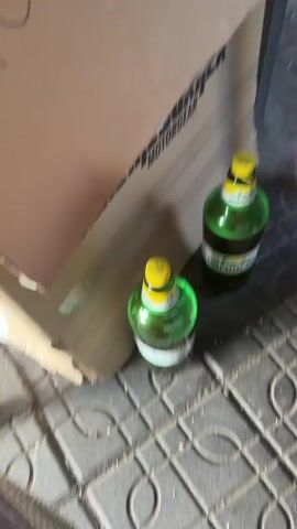 Two green bottles with yellow caps sit on gray patterned tile next to a cardboard box. A person's leg is visible on the right side of the frame.