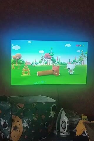 A television screen displays an animated scene. On the screen, two cartoon characters, one resembling a fox and the other a white bear, are sitting on a log. The fox character is holding a piece of wood. The background is a cartoon landscape with green grass, trees, and a blue sky with white clouds. The television is mounted on a wall, and below it are various objects, including a laundry basket with a space-themed pattern, an iron, and some yellow fabric. The screen has a colored light effect around its edges. The scene transitions to a different animated scene featuring a ladder and trees. Then, a new cartoon character, a bear wearing a top hat, glasses, and a bow tie, appears holding a wand.