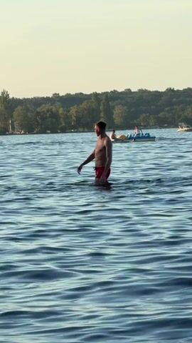 A man in red swim trunks stands in water up to his knees, facing away from the camera. He appears to be looking at something in the distance. In the background, several people are visible on a yellow inflatable float and on a blue pedal boat. The water is rippling, and there is a tree-lined shore in the distance. The sky is a pale, hazy blue, suggesting it is either early morning or late afternoon.