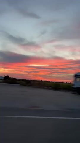 The video shows a truck driving on a road with a sunset in the background. The truck is white with a silver trailer and is moving from left to right across the screen. The sunset is colorful, with shades of orange, pink, and purple. The sky is cloudy, and the clouds are illuminated by the setting sun. The road is paved, and there are white lines marking the lanes. There are some trees and fields visible on the side of the road. The video is shot from a moving vehicle, which creates a sense of motion blur.