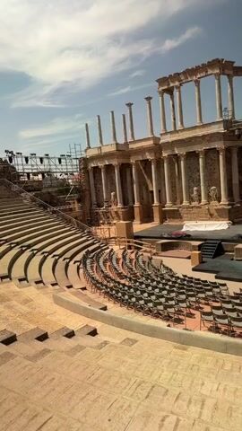 The video shows a wide-angle view of an ancient Roman theater. In the foreground, rows of empty seats are visible, made of stone and arranged in a semi-circle. The seats are facing a stage area. The stage itself appears to be set up for a performance, with black fabric covering parts of it and a theatrical prop resembling a stylized tree or figure with dark fabric draped over it. Behind the stage, the remains of the theater's impressive architecture are visible. This includes a grand facade with multiple levels of columns, arched openings, and stone stonework. Some of the upper parts of the structure appear to be in ruins. In the background, scaffolding and stage lighting equipment are set up, suggesting preparations for an event. The sky above is blue with some scattered white clouds. The overall scene is bathed in sunlight, indicating it is daytime. The video pans slightly to the right, offering a broader view of the seating area and the stage. There are no people actively performing or interacting on the stage. A few individuals are visible in the background, near the architectural structure, possibly workers or staff involved in setting up.